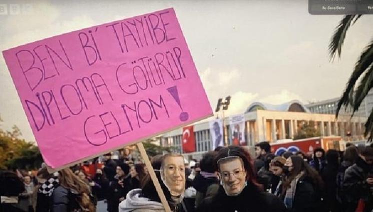 Étudiante masquée brandissant une pancarte ironique sur l’attribution d’un diplôme à un dirigeant