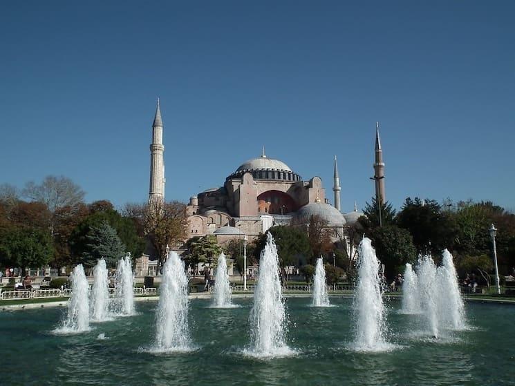 Vue extérieure de la mosquée Sainte-Sophie à Istanbul, avec ses minarets majestueux et une fontaine au premier plan sous un ciel bleu dégagé.
