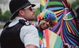 Un policier flouté au premier plan, et derrière lui un homme souriant, les cheveux et le maquillage aux couleurs de l'arc-en-ciel. 